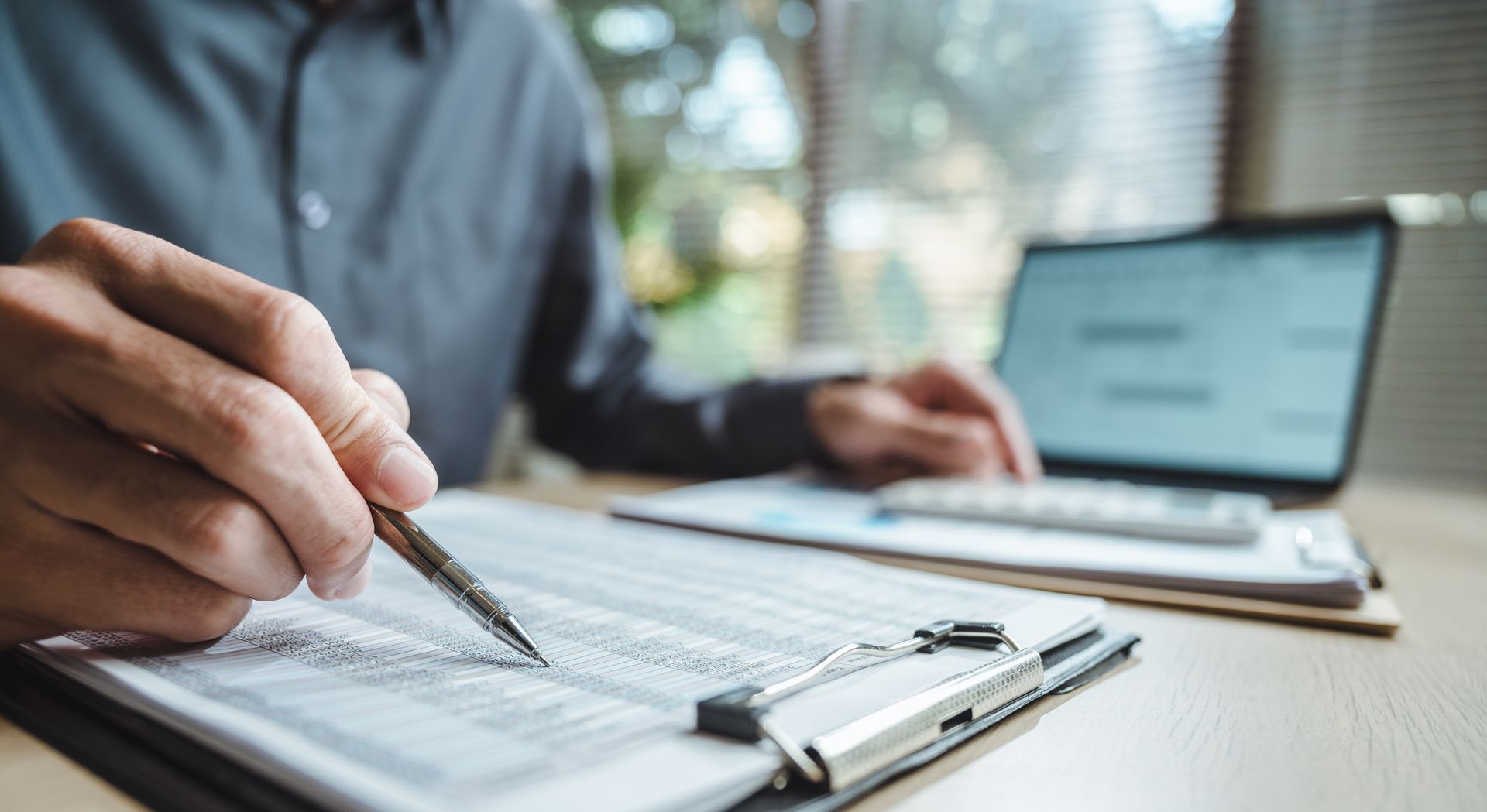 Person in grey shirt reviews financial documents, using a pen and calculator near a laptop.