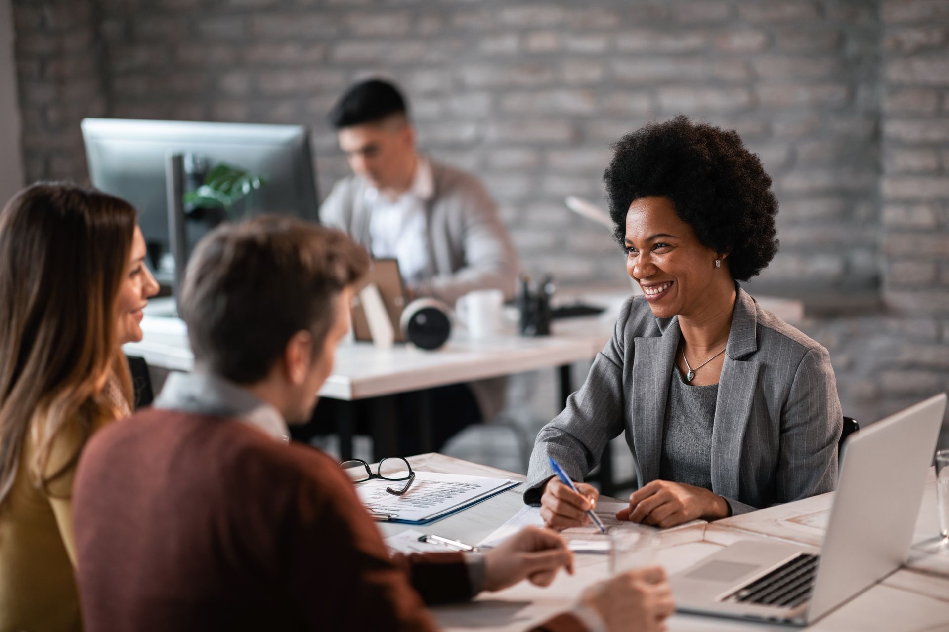 Woman in gray blazer smiles, talking to couple at a table, another person at a desk in background. Office setting.