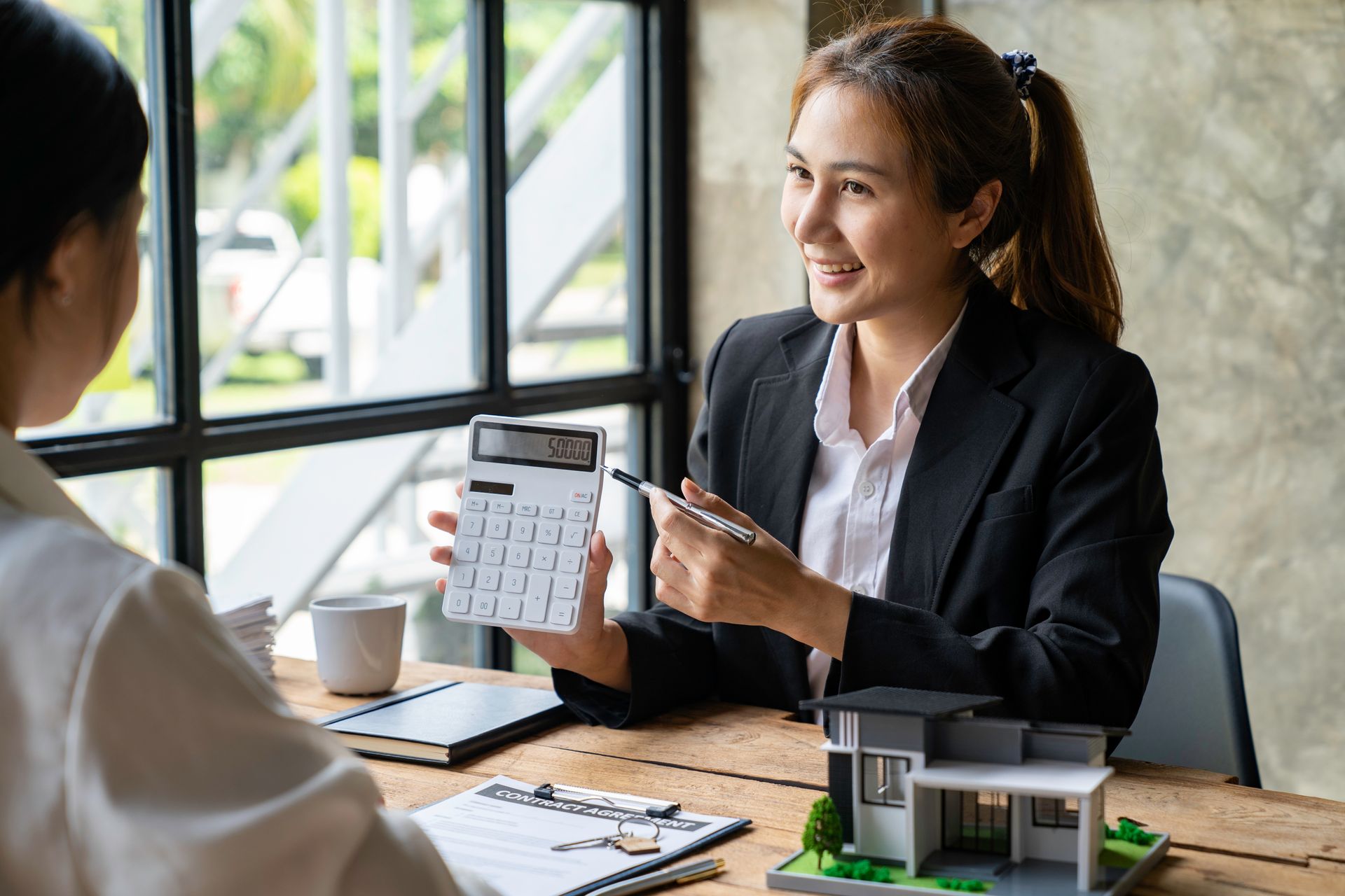 A real estate agent in a suit shows a calculator to a client, model house on desk, natural light.