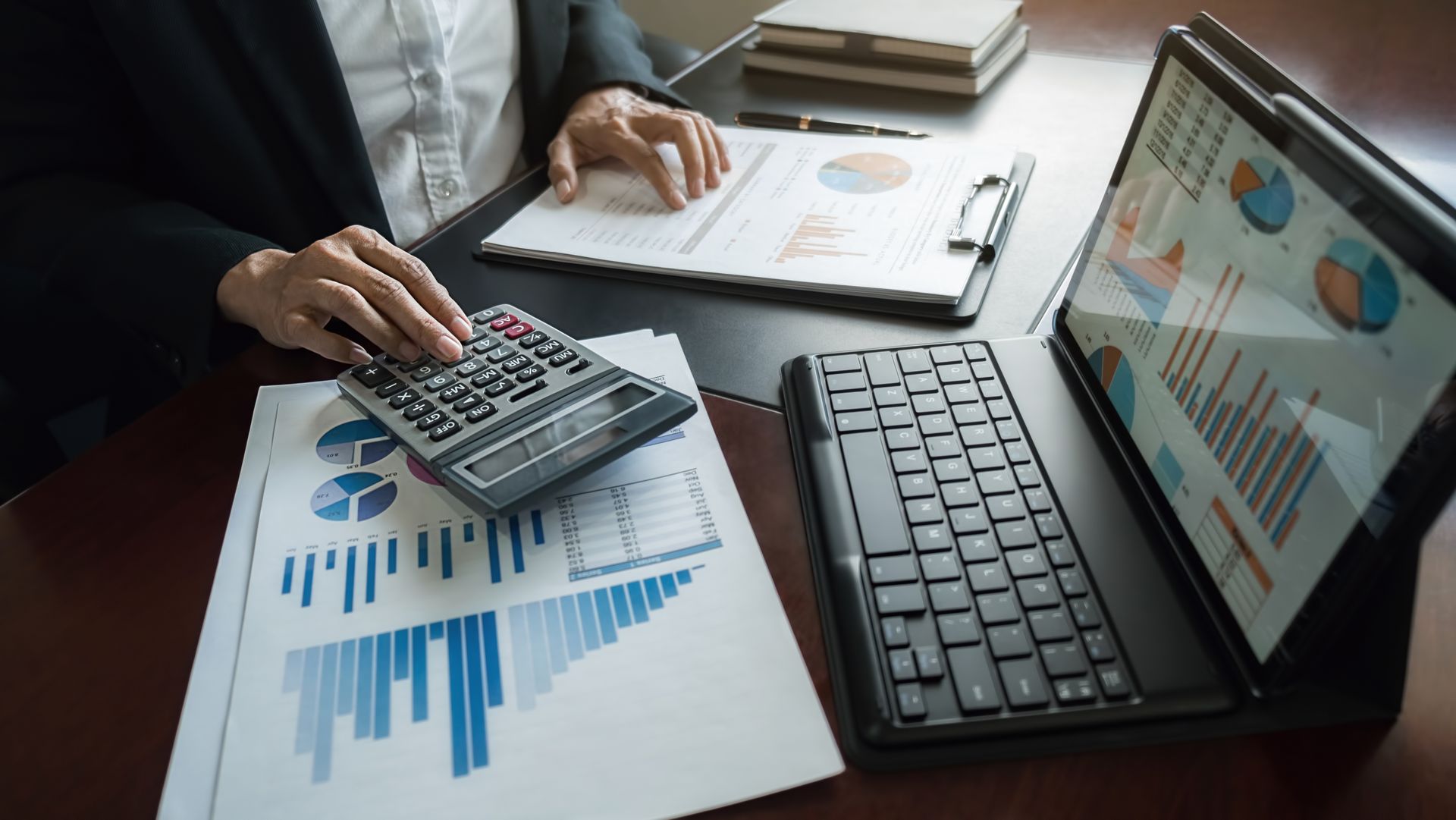 Person calculating with calculator, surrounded by charts and graphs, with laptop on table.