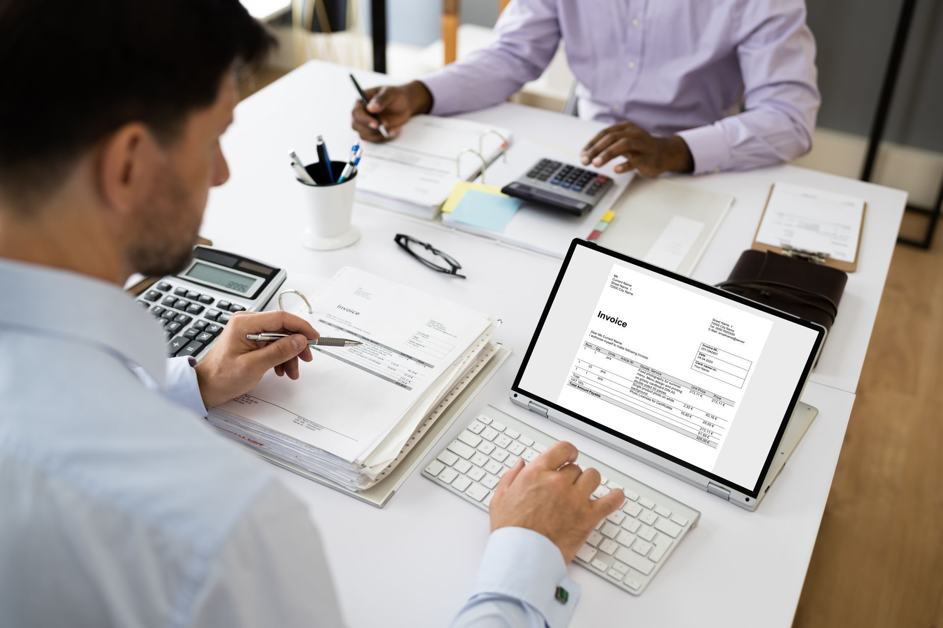 Two accountants working on invoices at a desk; one on a computer, the other writing.