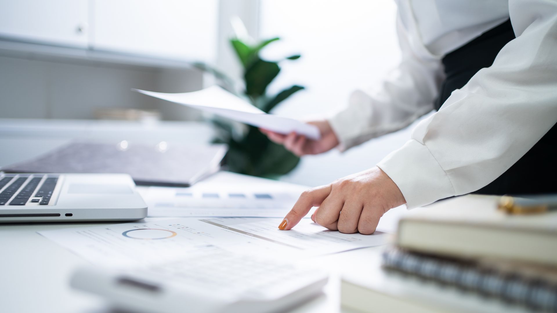 Person pointing at a chart while holding papers near a laptop in an office setting.