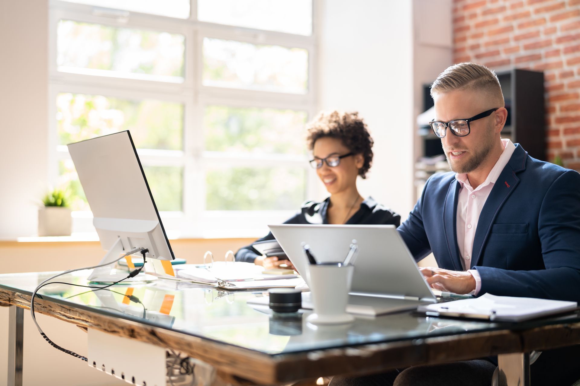 Two colleagues working together at a desk, one using a computer and the other a laptop, in an office setting.