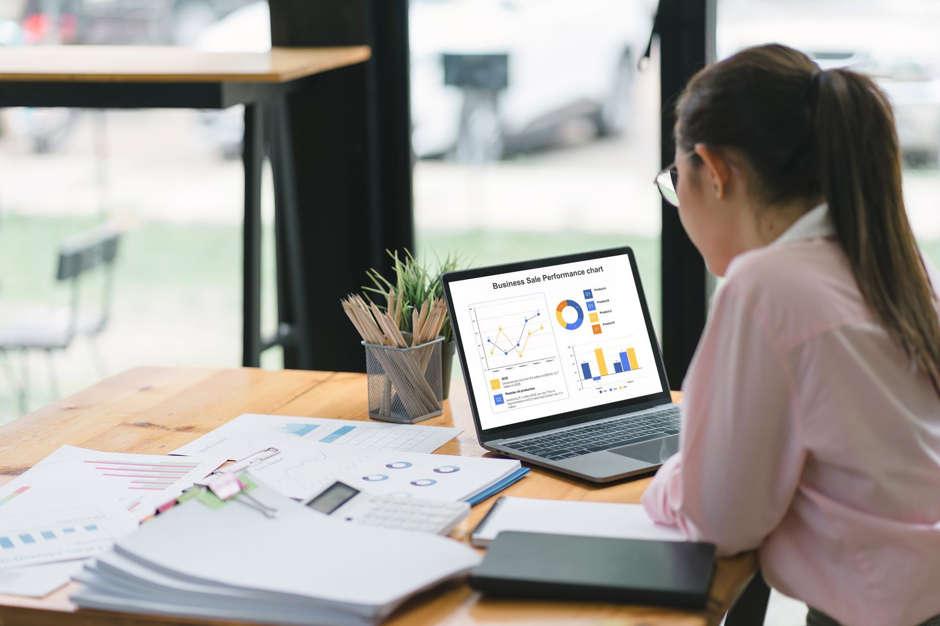 Woman working on laptop, viewing charts and graphs, surrounded by documents and calculator at a wooden desk.