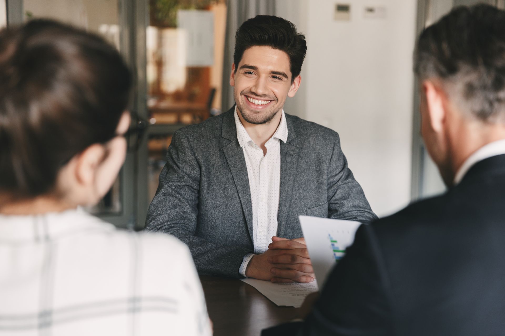 Man in a blazer smiling, sitting at a table across from two people, in an office.