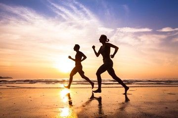 Two people running on a beach at sunset, silhouetted against the sky.