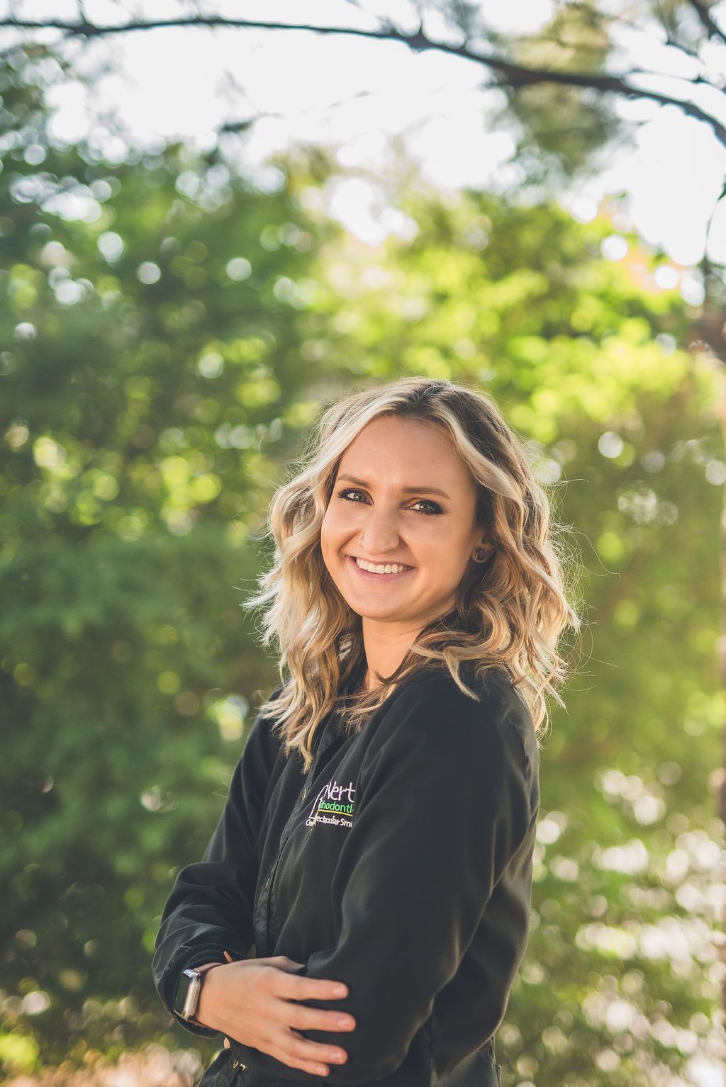 a woman in a black jacket is standing in front of trees with her arms crossed .