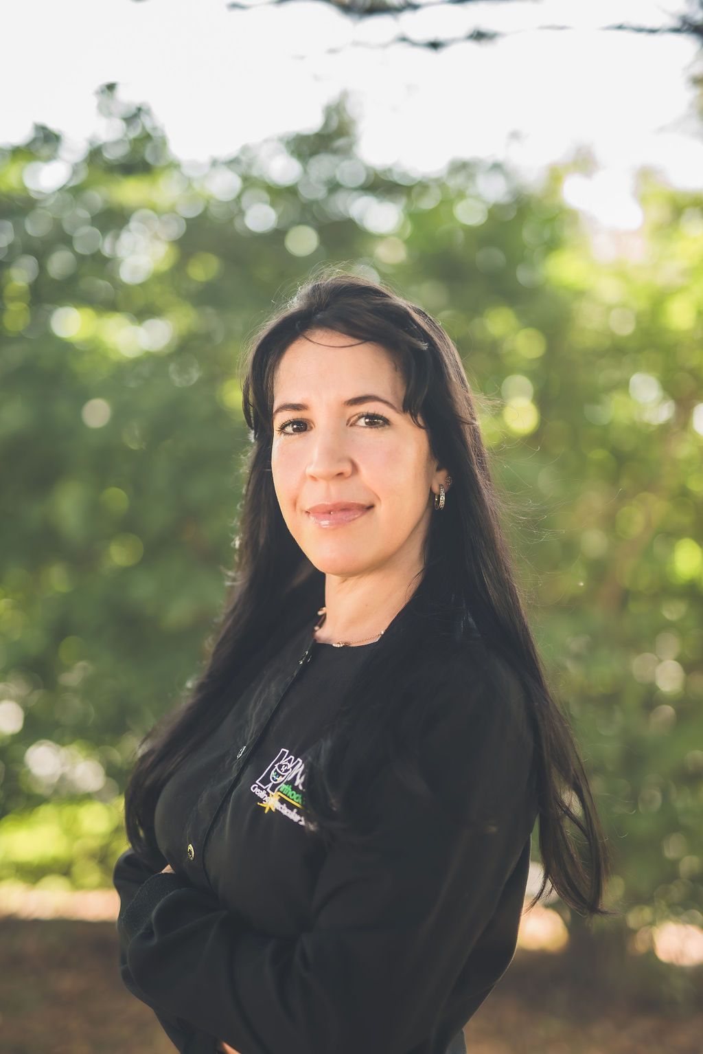 a woman in a black jacket is standing with her arms crossed in front of trees .
