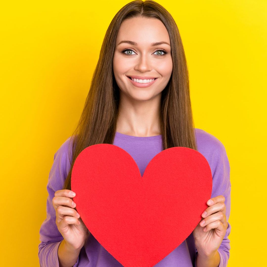 A woman is holding a red heart in her hands on a yellow background.