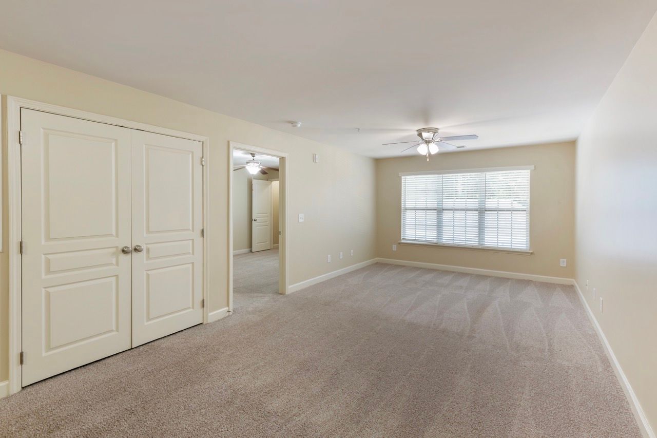 Empty beige living room with window, blinds, ceiling fan, and double closet doors.