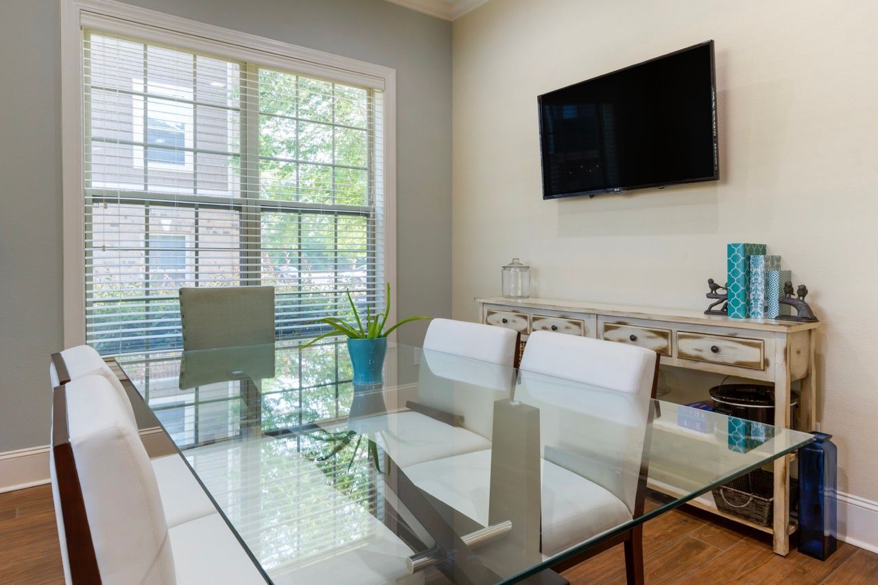 Dining area in an apartment with a glass table, white chairs, a large window, and a wall-mounted TV.