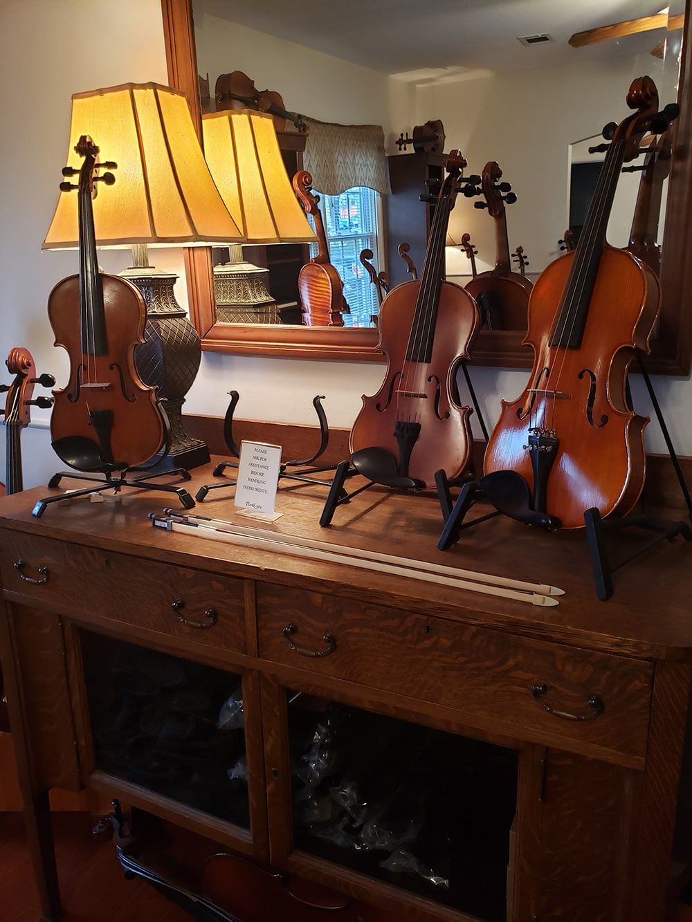 Three violins displayed on stands atop a wooden sideboard, with two table lamps positioned between them.