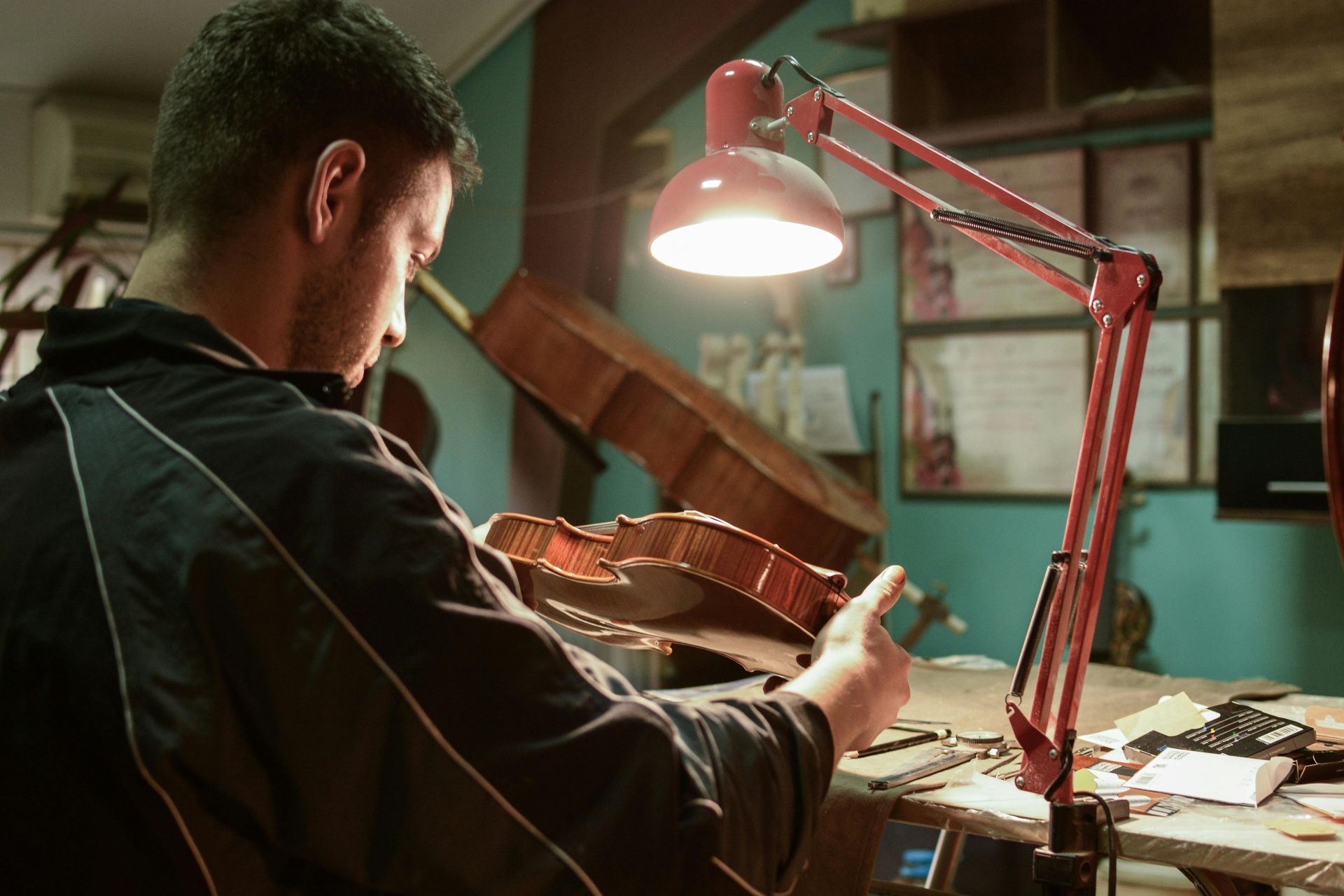 A person examines a wooden violin body under the focused light of a desk lamp in a workshop.