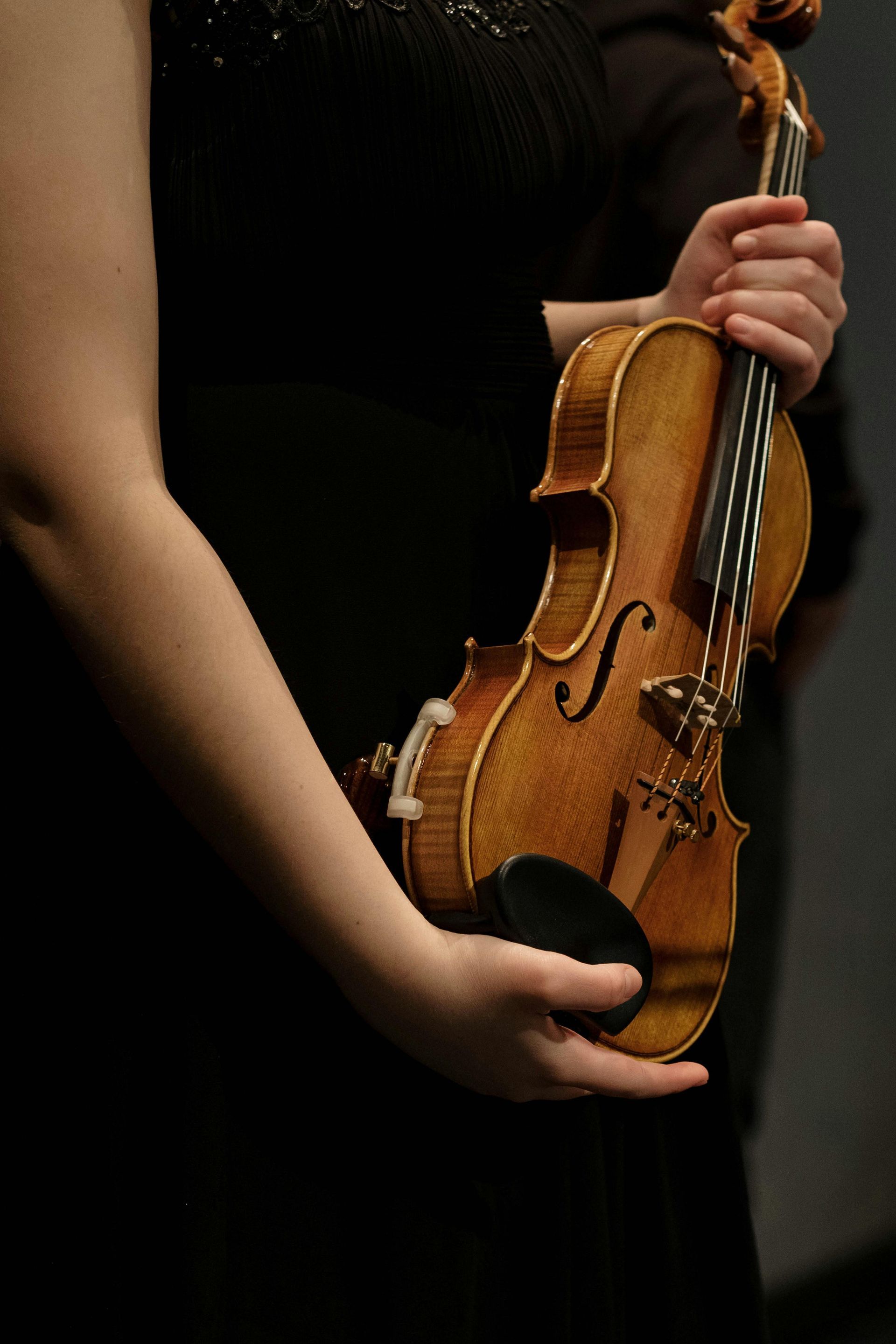A person in a black gown holds a wooden violin against their torso.