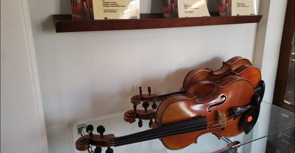 Two violins resting on a glass display table beneath a wooden shelf with information cards.
