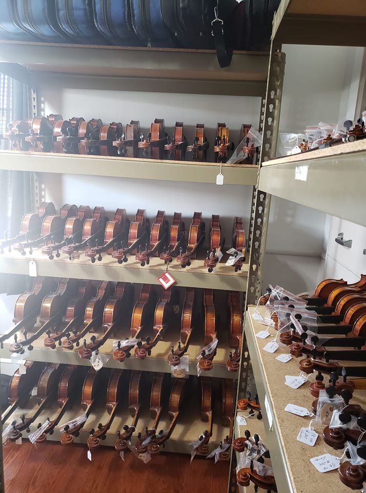 A warehouse shelf stocked with rows of wooden violins, many with white price tags attached to the necks.