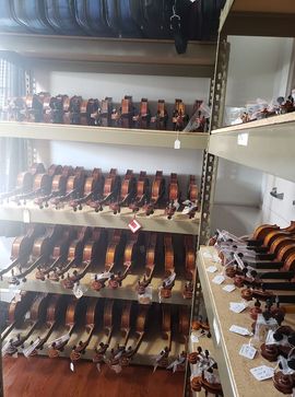 A warehouse shelf stocked with rows of wooden violins, many with white price tags attached to the necks.