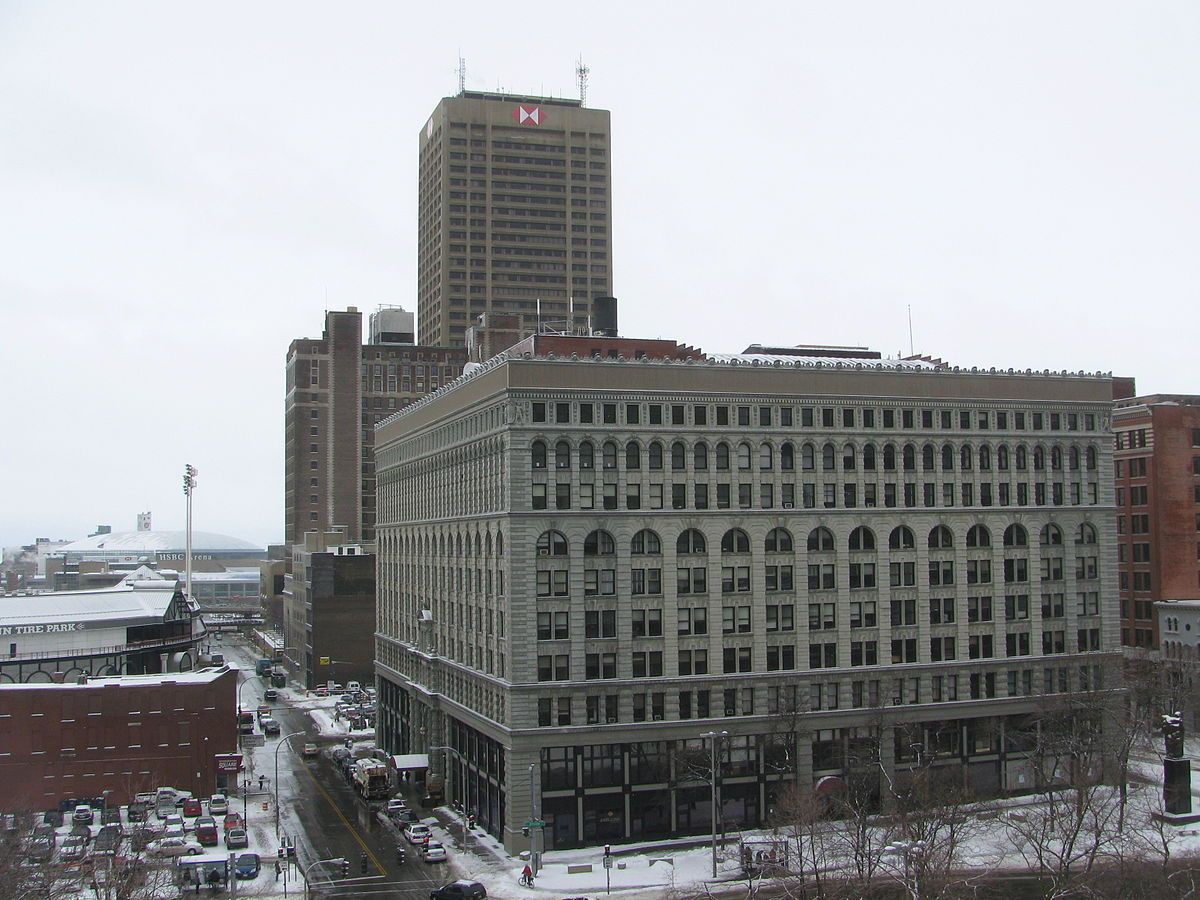 A large stone building with arched windows stands in a snowy cityscape next to a taller modern tower.
