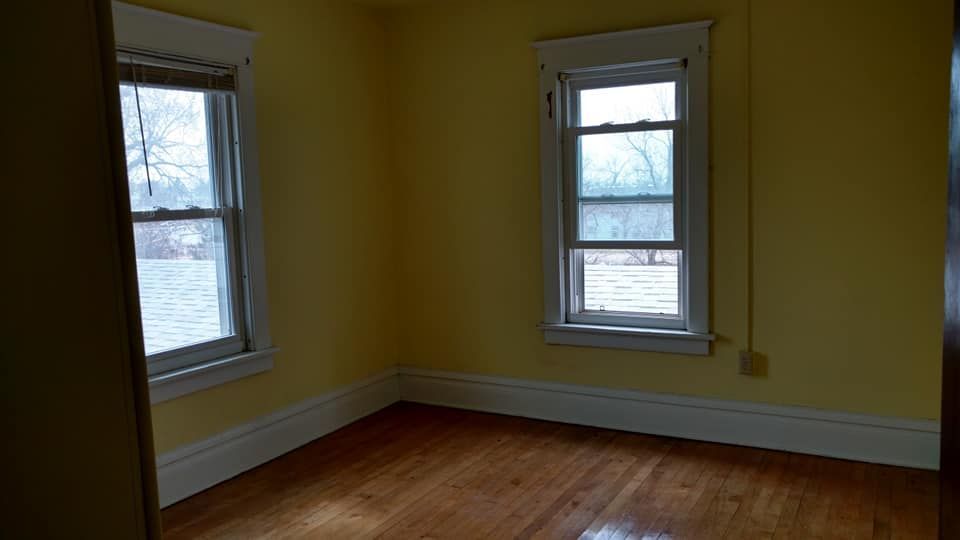 An empty room with yellow walls and hardwood floors and two windows.