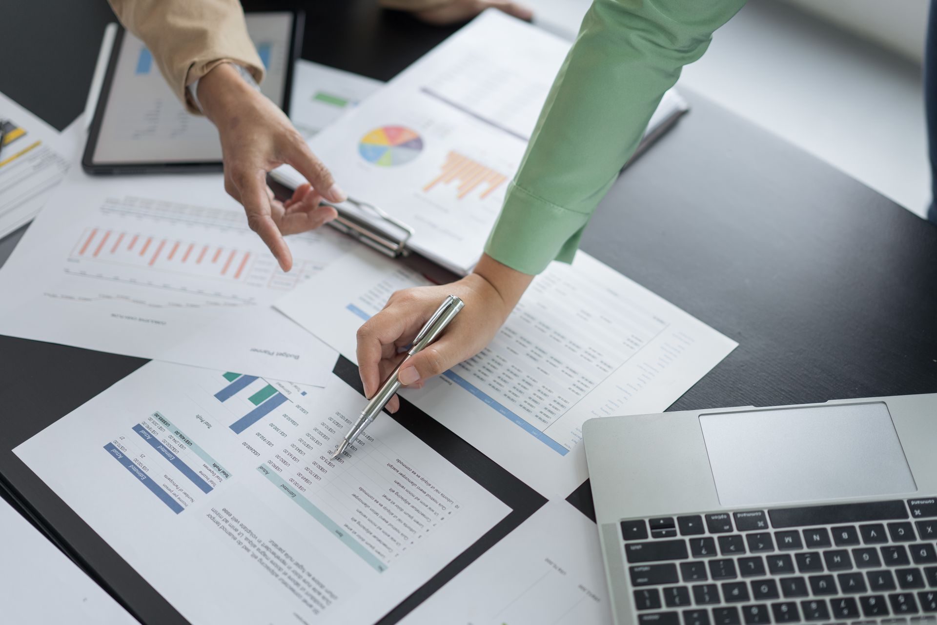 Two people examining charts and graphs on a table with a laptop and tablet, pointing to data Two people examining charts and graphs on a table with a laptop and tablet, pointing to data