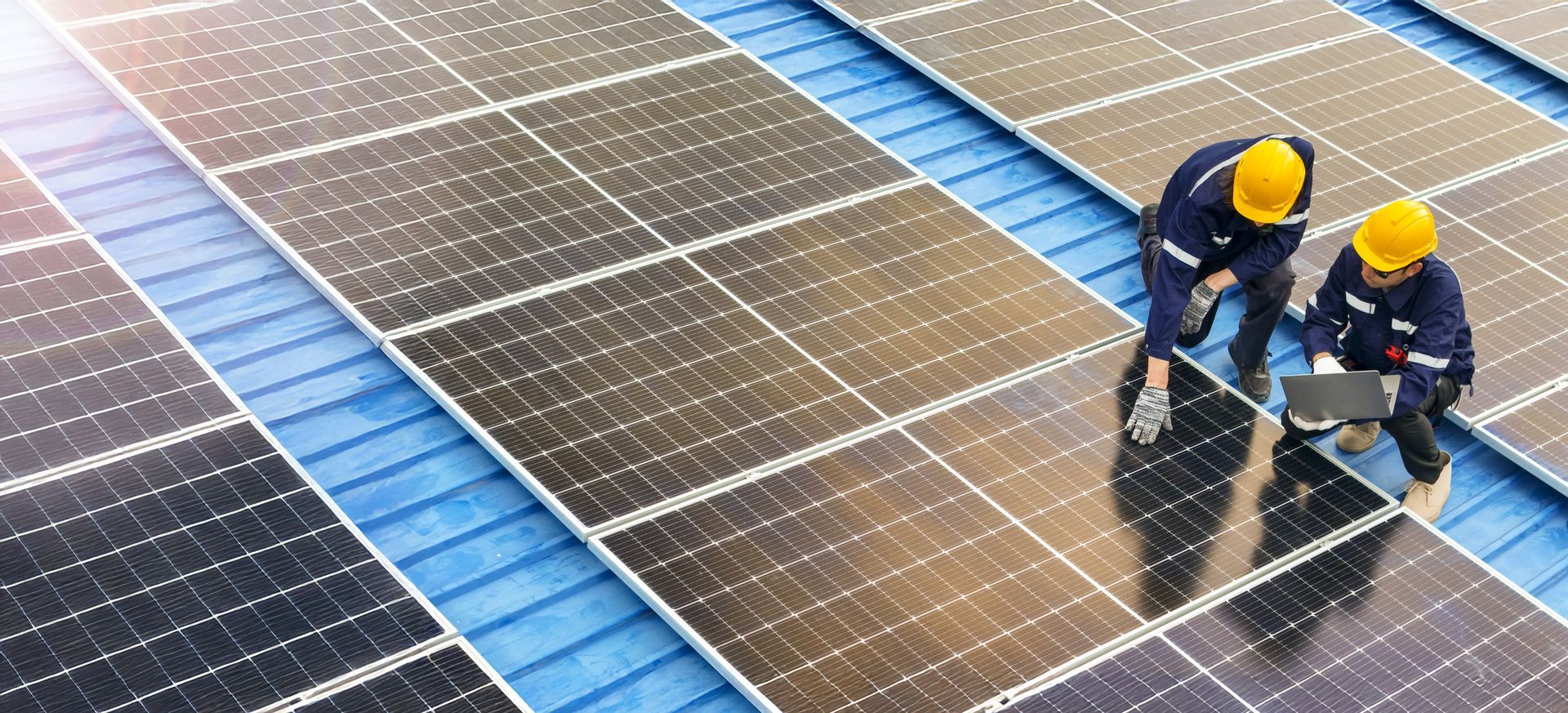 Two workers in yellow helmets inspect solar panels on a blue roof