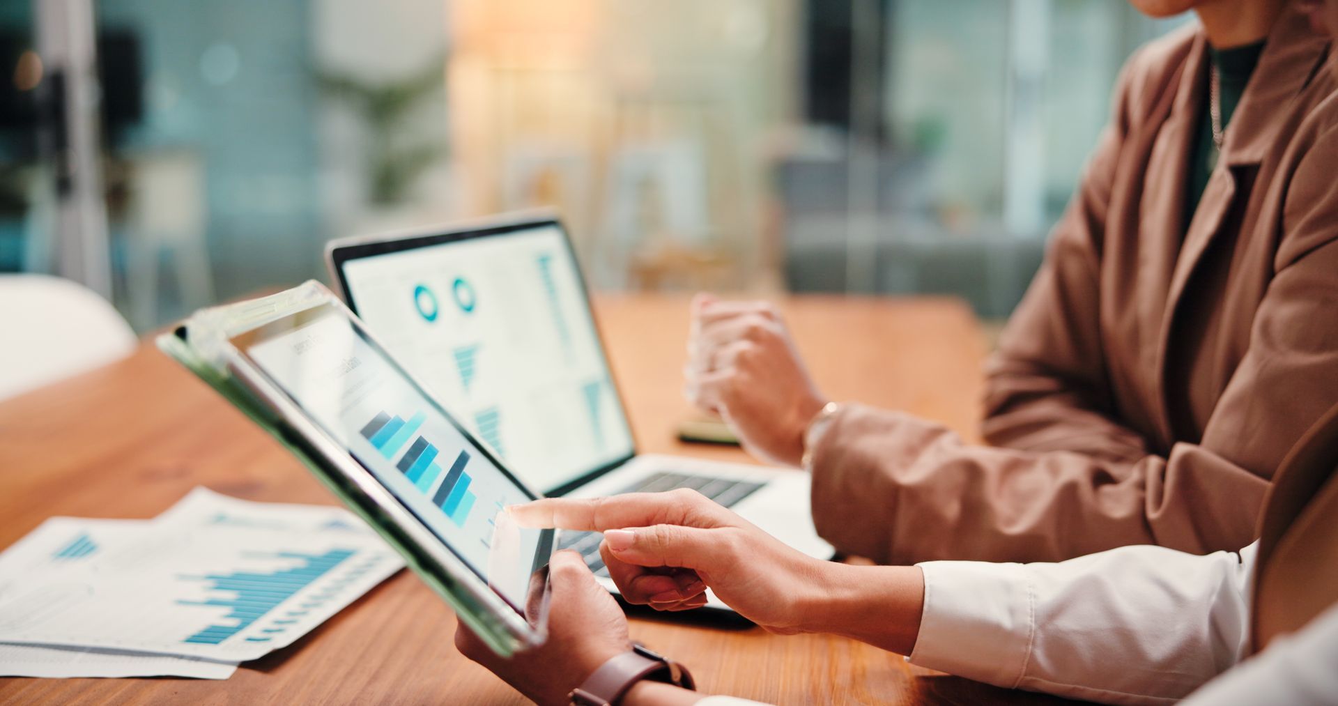 Two people analyze charts on a tablet and laptop at a wooden table in an office