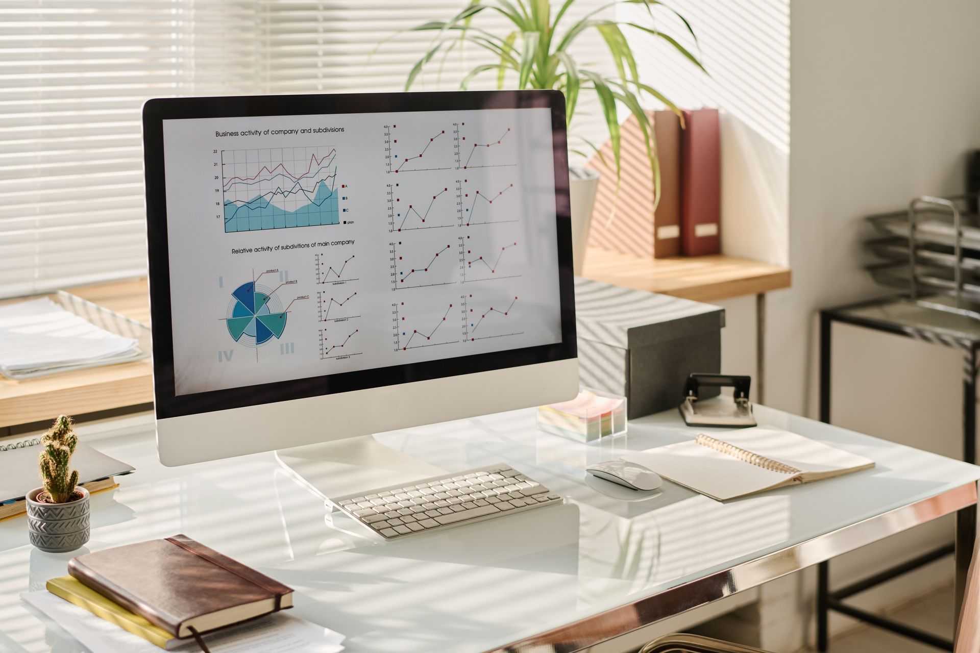 Desktop computer showing charts, on a modern desk with office supplies and a plant, lit by sunlight