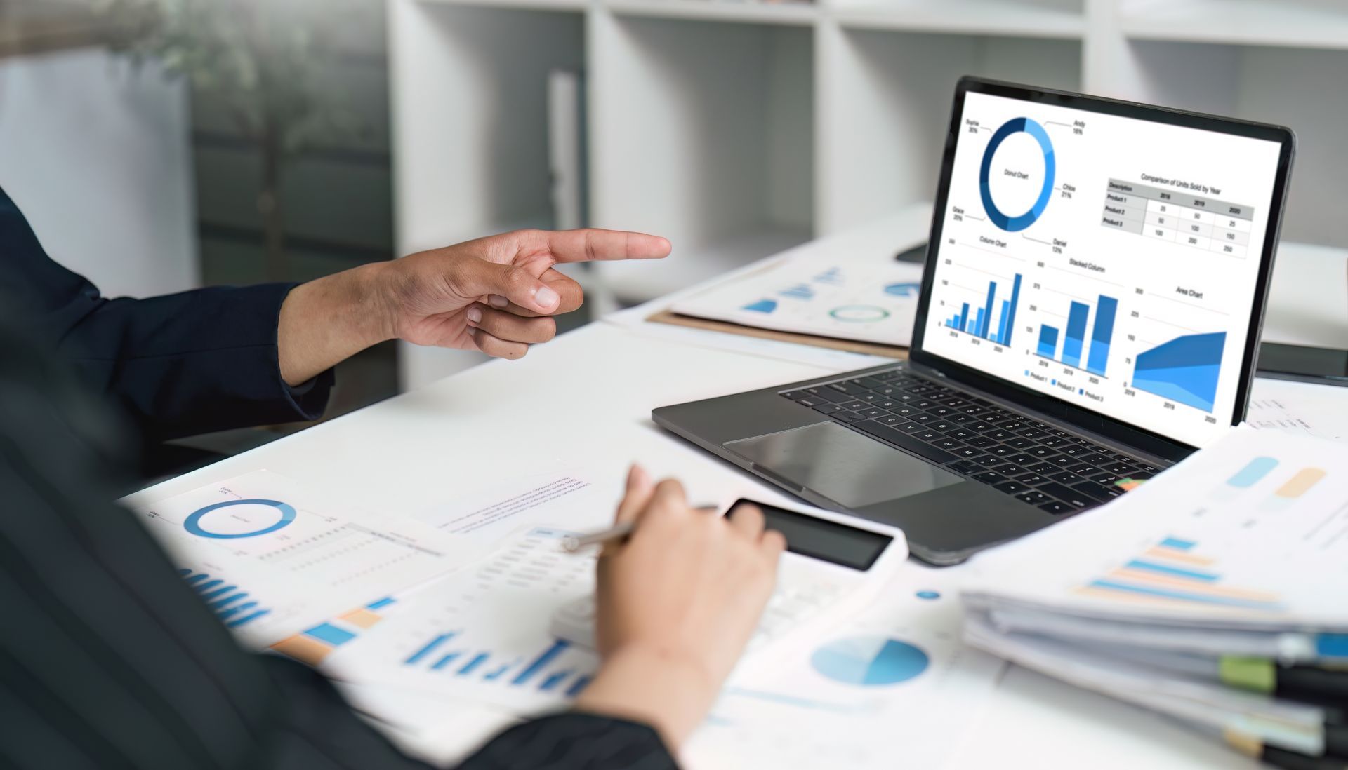 Person points at a laptop displaying financial data, using a calculator amid charts and documents