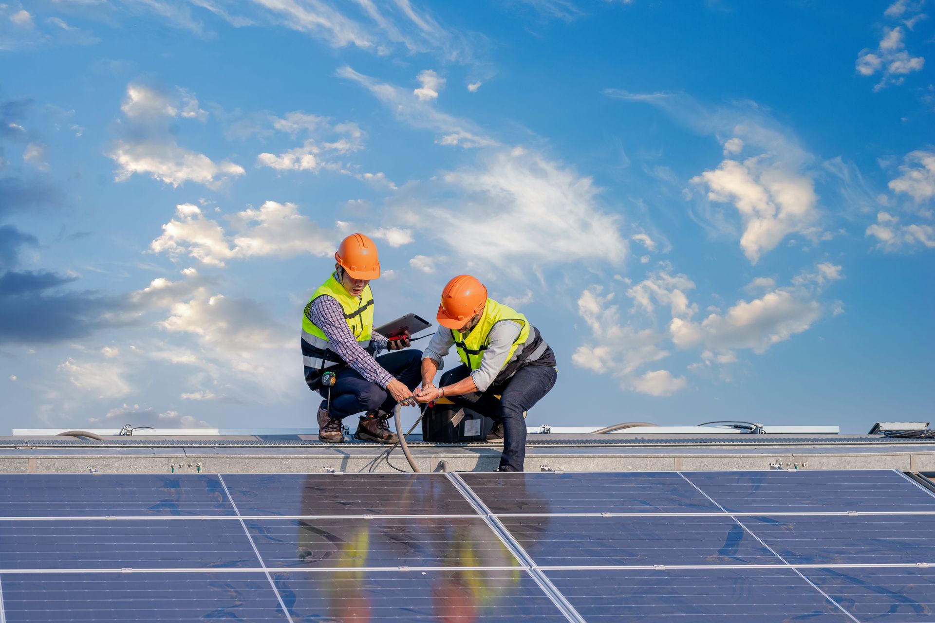 Two workers in orange helmets install solar panels on a roof, blue sky above