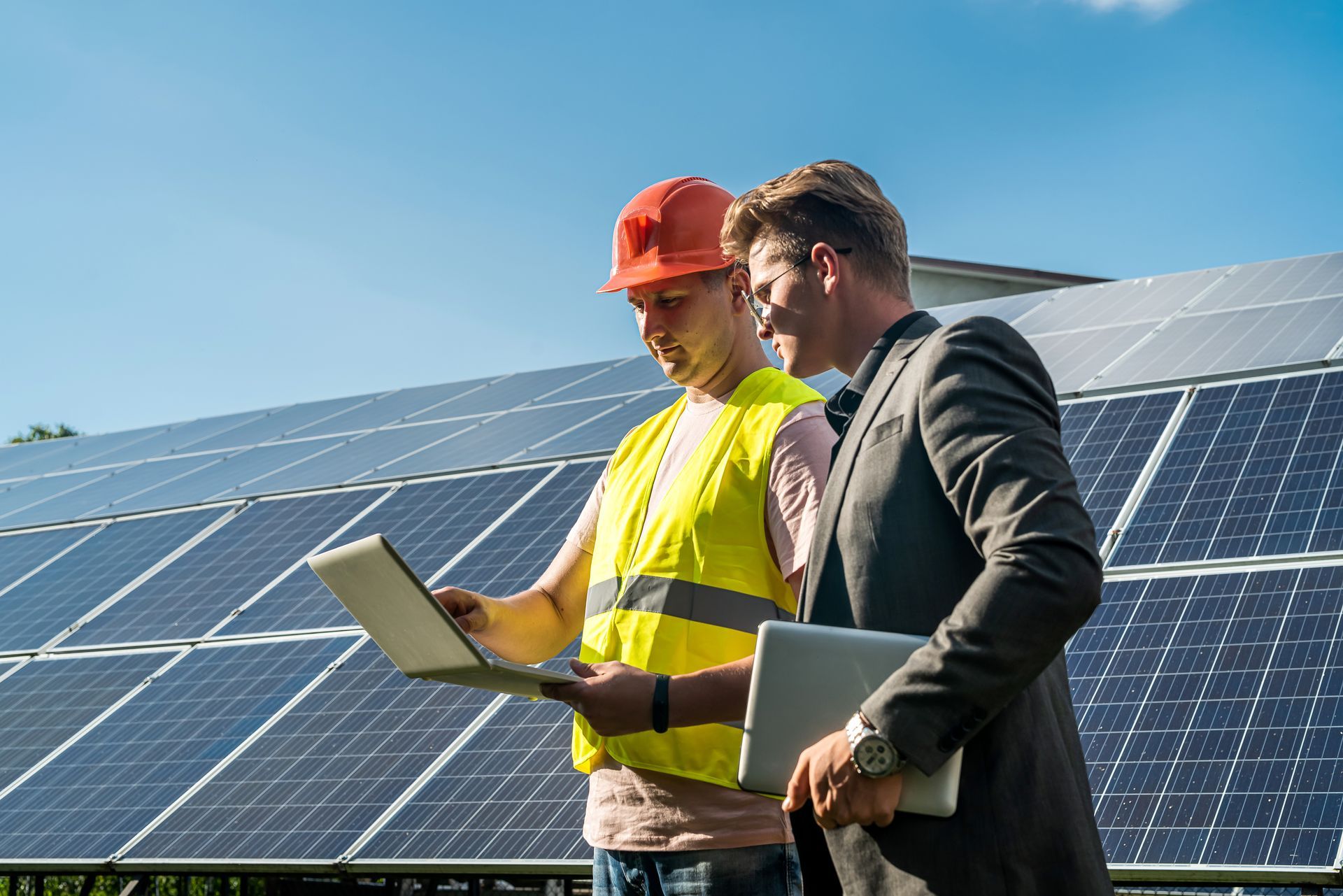Two men look at a laptop in front of solar panels Two men look at a laptop in front of solar panels