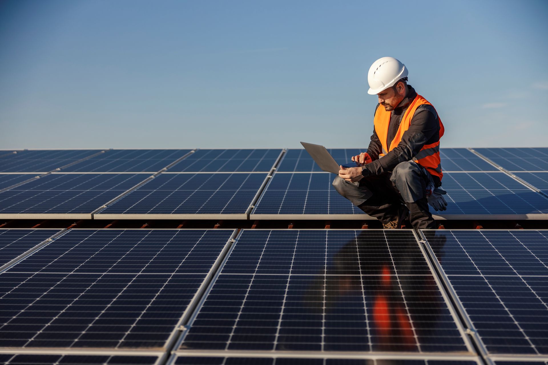 Man in safety vest and hard hat working on a laptop atop solar panels