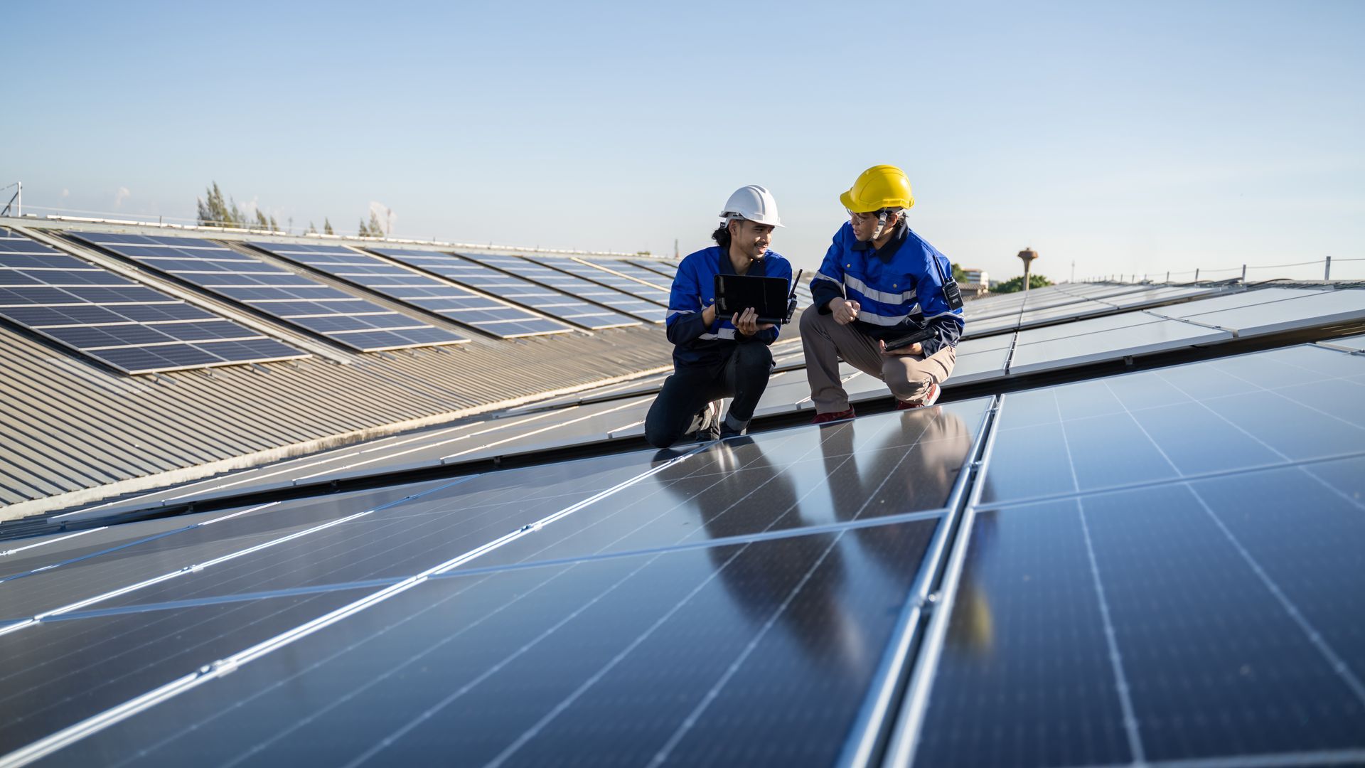 Two workers, one with a yellow hard hat and another with a white hard hat, inspecting solar panels on a rooftop