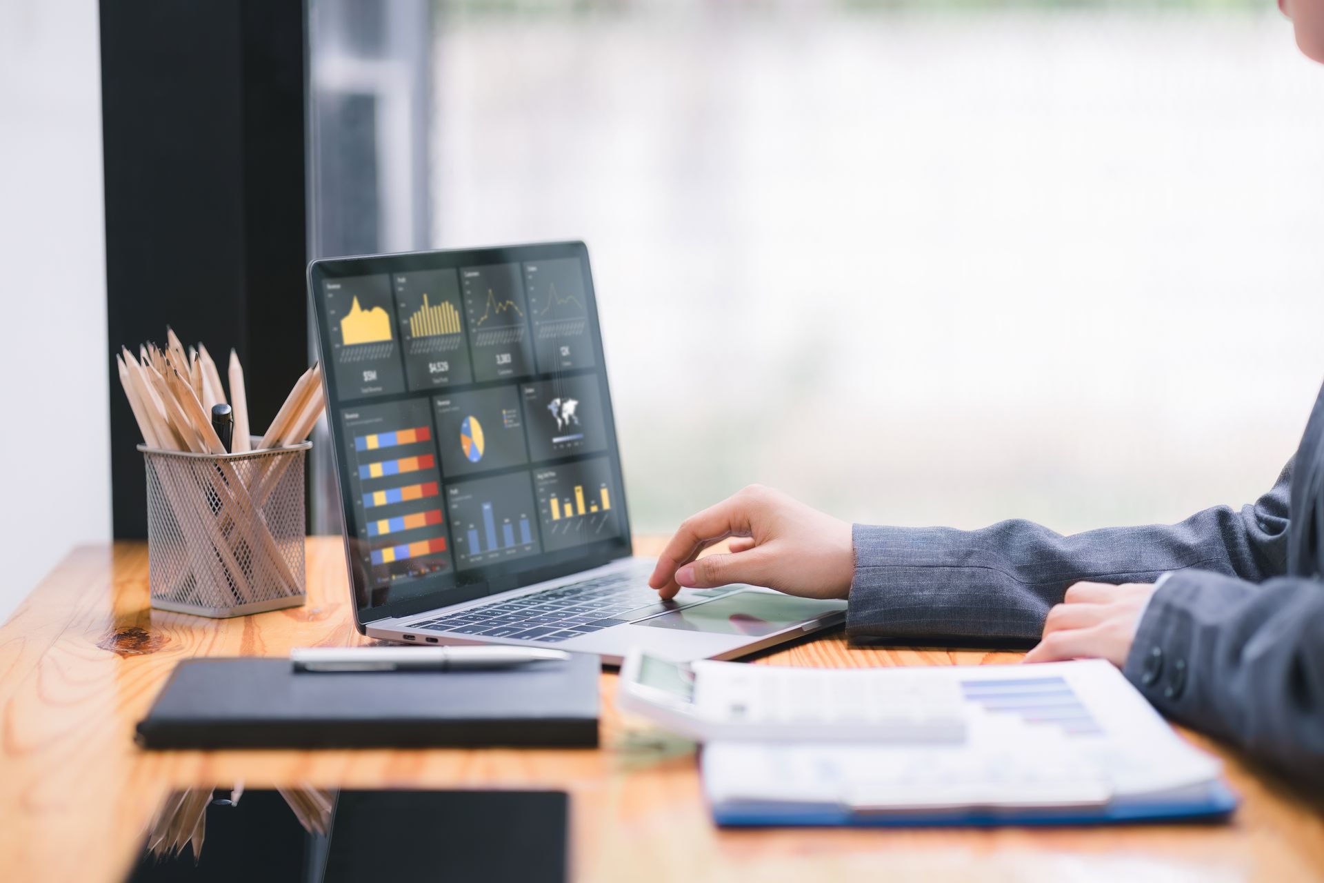 Person using a laptop displaying financial charts and data, with papers, calculator, and pencils on a desk