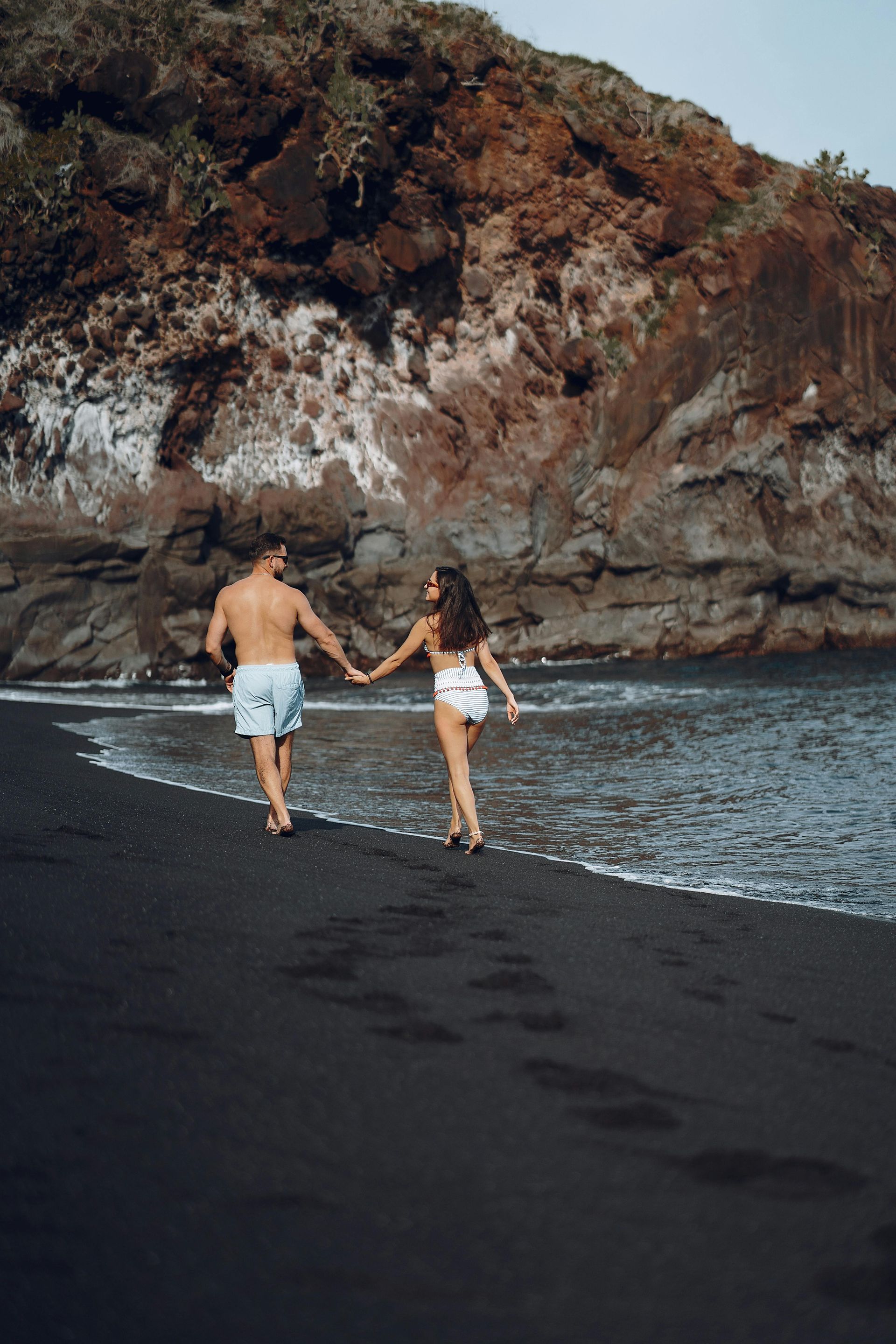 A man and a woman are walking on a black sandy beach holding hands.