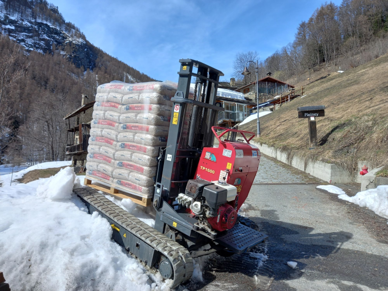 Un carrello elevatore cingolato rosso trasporta un pallet di sacchi di cemento in salita su un sentiero di montagna innevato.