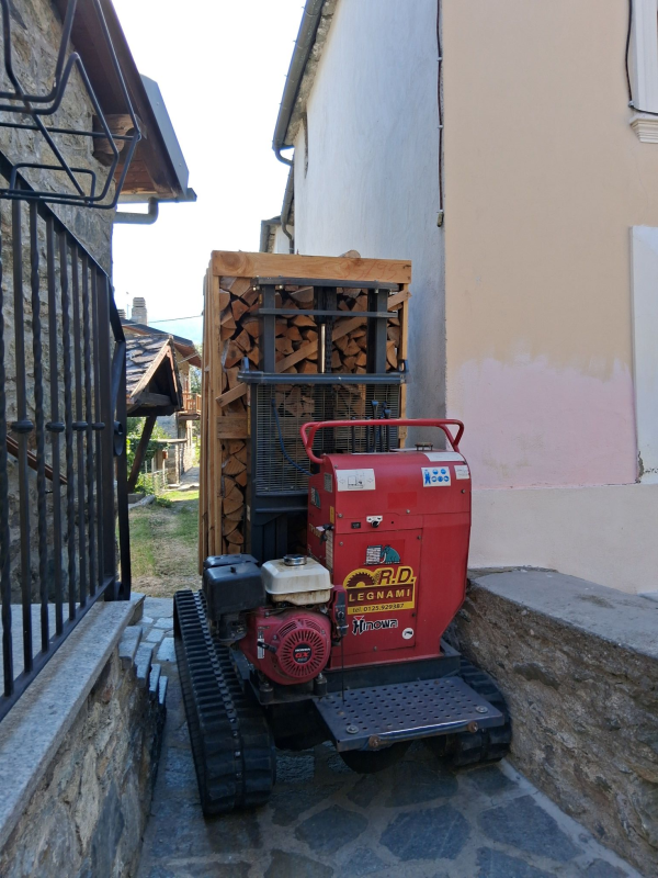 Un veicolo cingolato rosso trasporta un pallet di legna da ardere accatastata attraverso uno stretto vicolo fiancheggiato da muri in pietra.