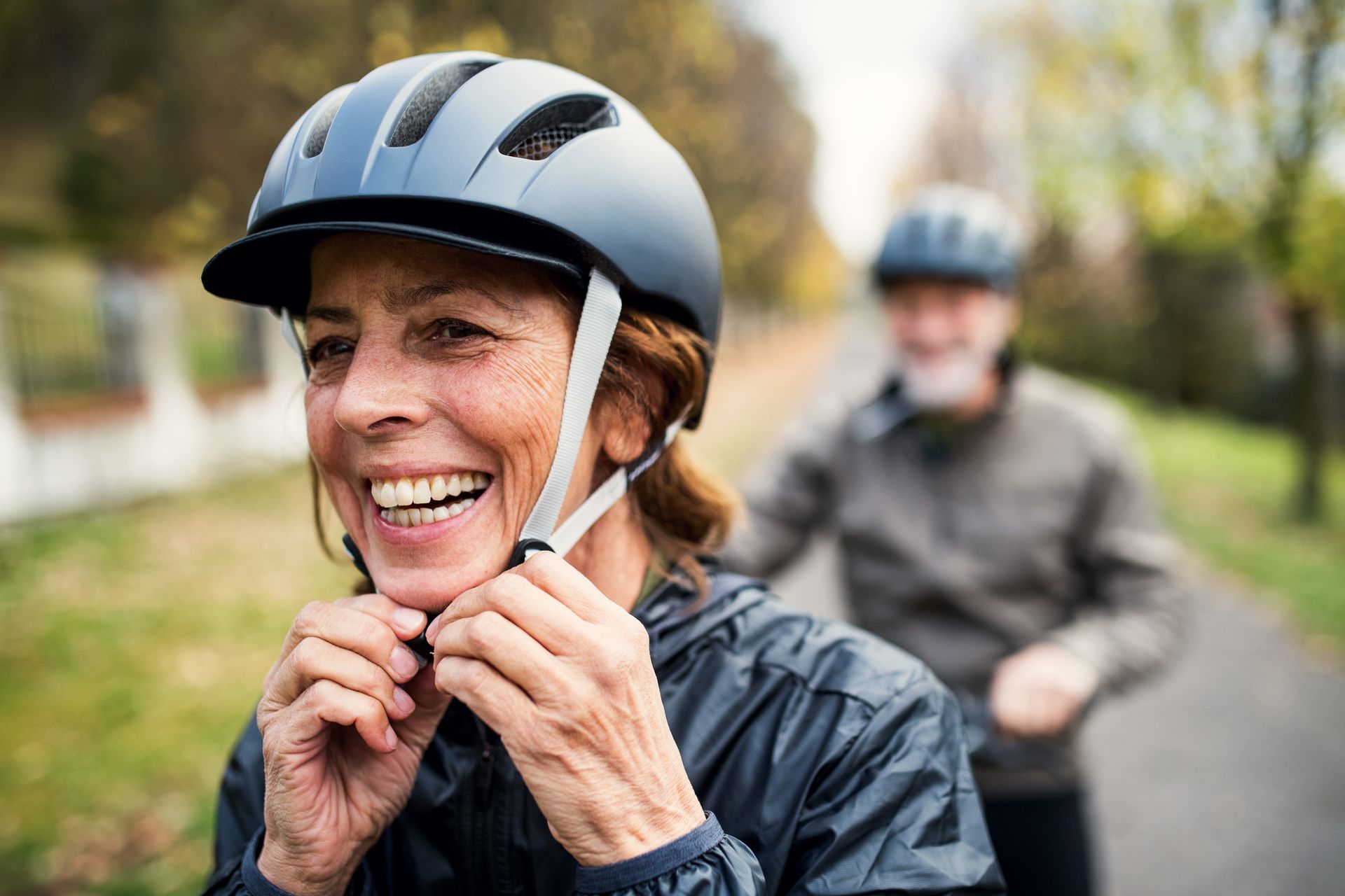 Woman strapping on a bicycle helmet with man on a bike behind her wearing a helmet.