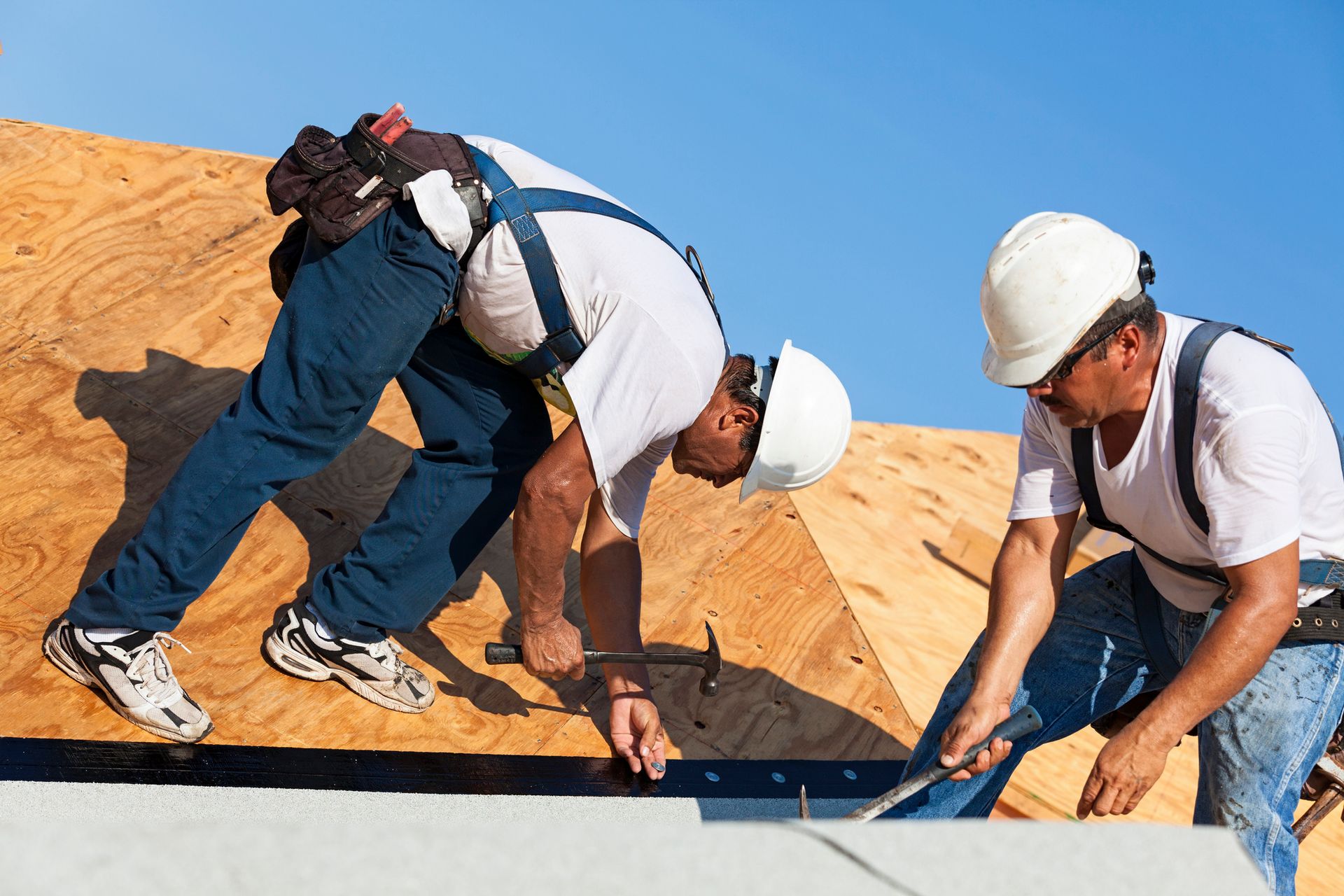 Two construction workers wearing safety gear installing roofing materials with hammers Two construction workers wearing safety gear installing roofing materials with hammers