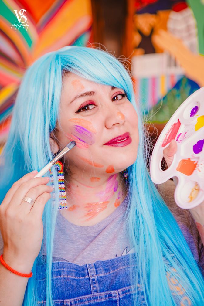Woman with blue wig covered in paint, holding palette and paintbrush, smiling.