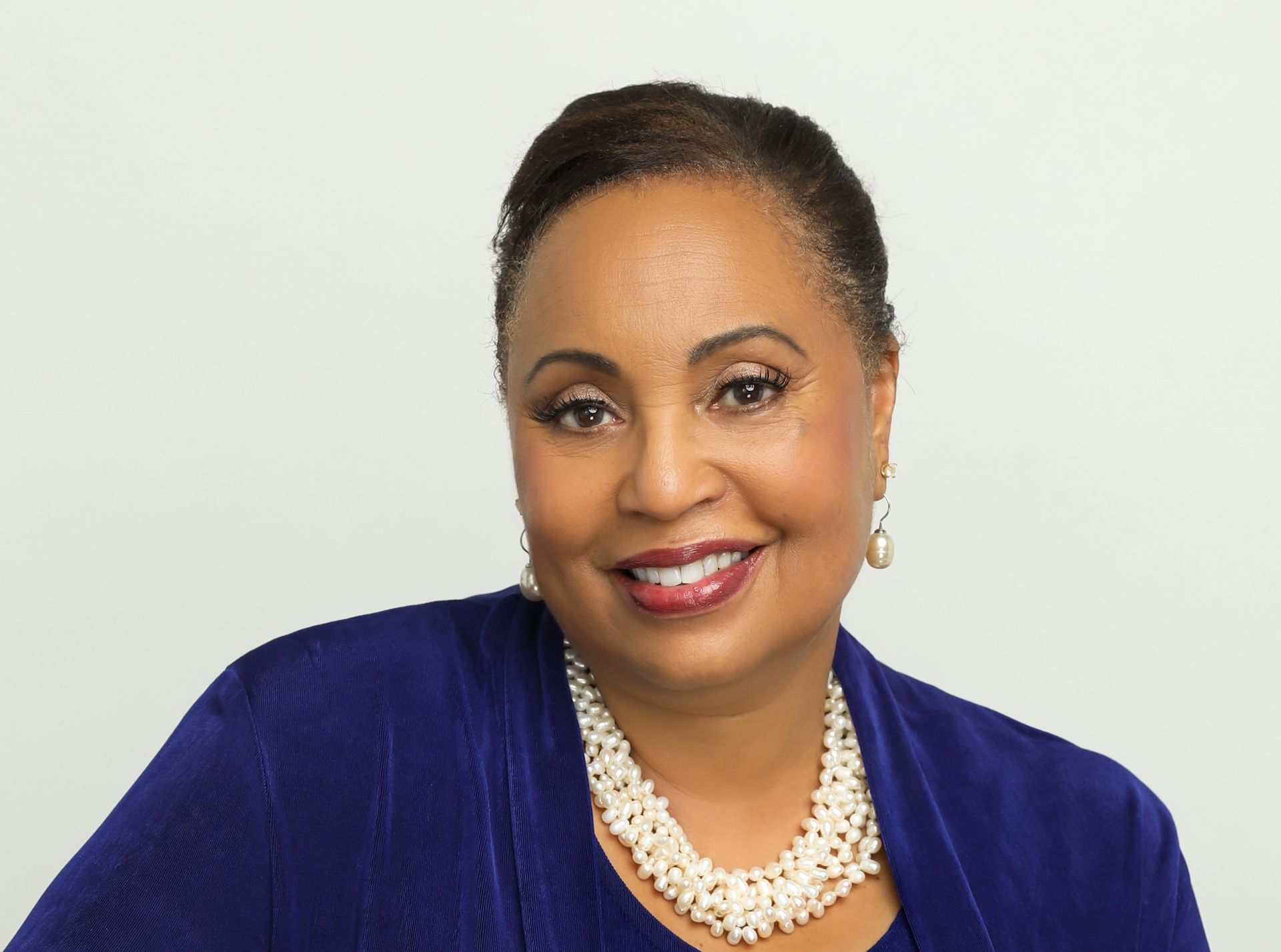 Woman in blue top, smiling, wearing pearl necklace, against a white background.
