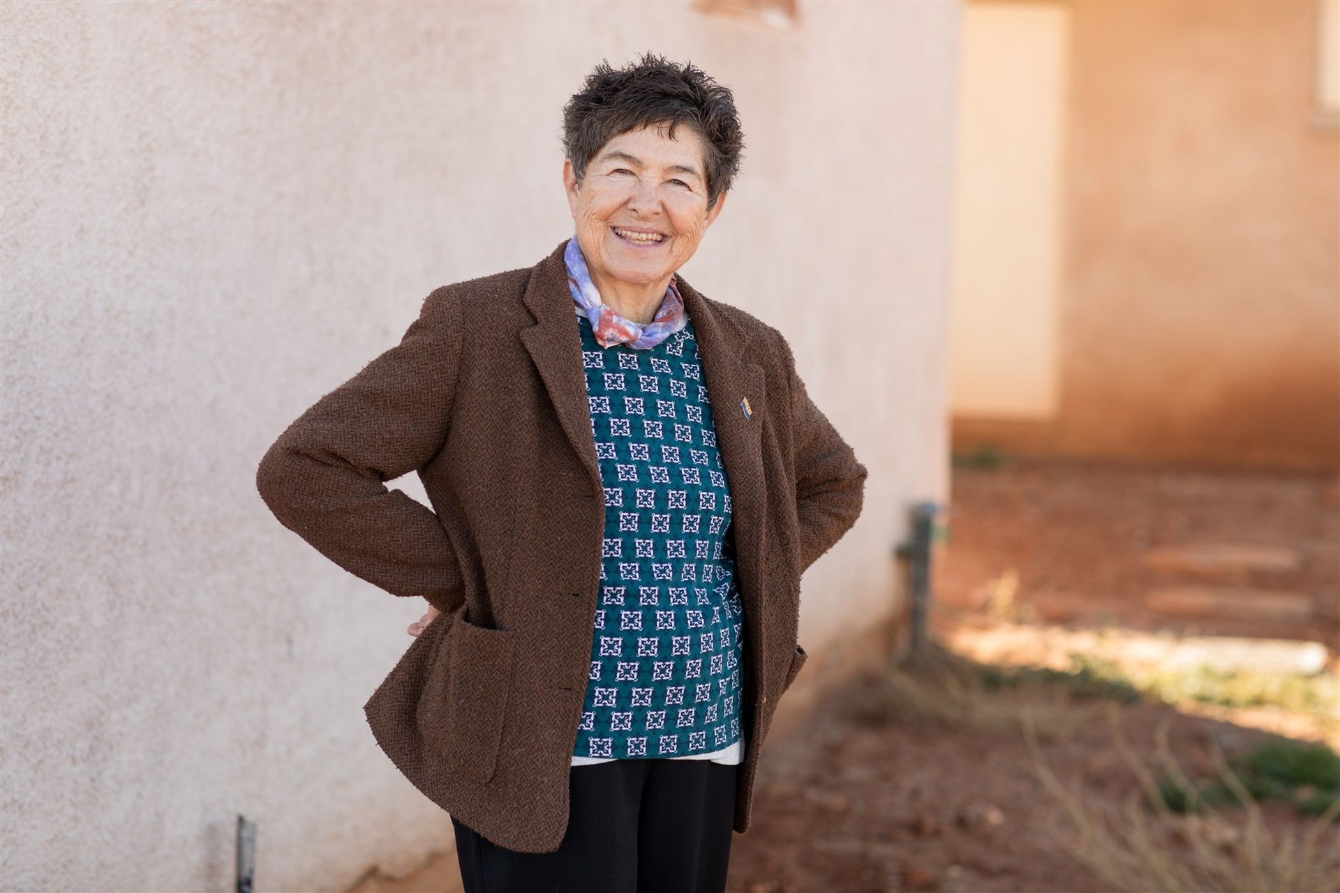 Woman with short, dark, spiky hair and glasses, smiling, wearing a purple shirt, beaded necklace, sitting on a couch.