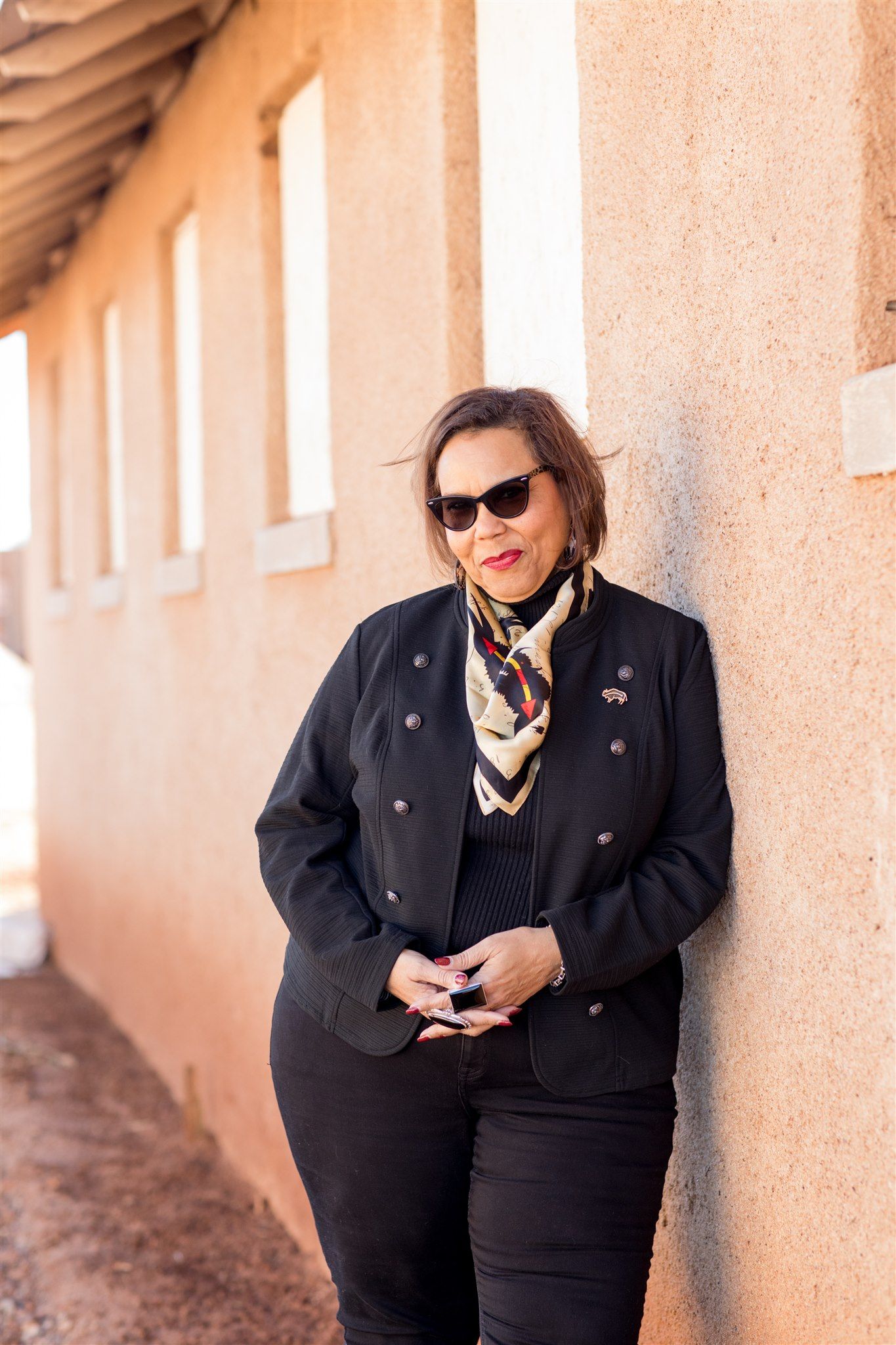 Woman in sunglasses and black jacket leans against a beige wall, a scarf around her neck.
