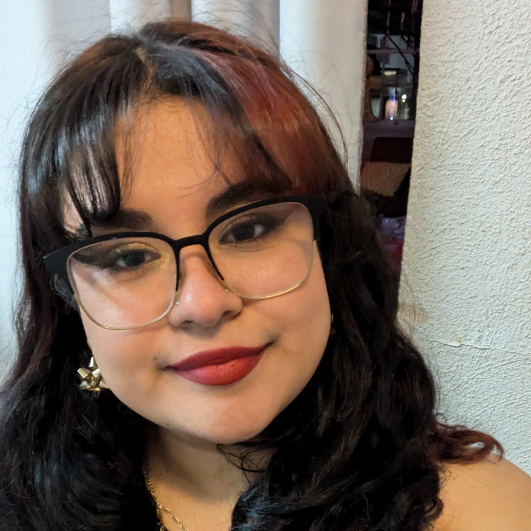 Woman with glasses, red lipstick, and dark curly hair; a close-up shot indoors, looking at the camera.