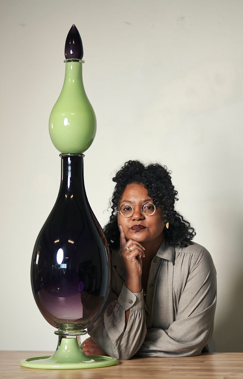 Woman poses with large, layered glass sculpture. Green and purple hues. Wooden table.