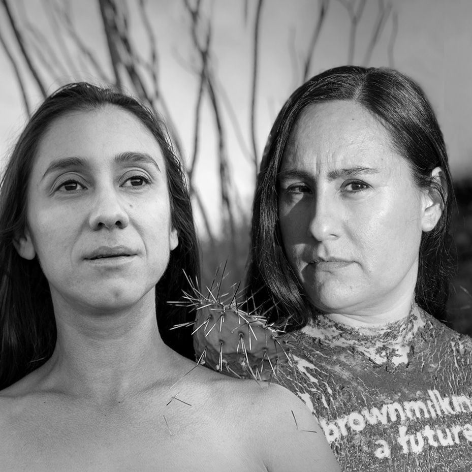 Two women, facing forward. One is bare-chested; the other has a t-shirt. Both have serious expressions.