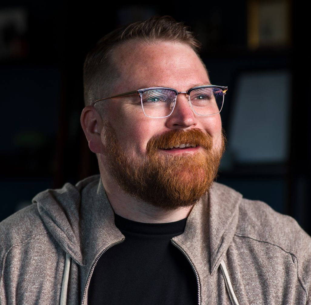 Man with a red beard, glasses, and a gray hoodie smiles, looking slightly off-camera in a dark room.