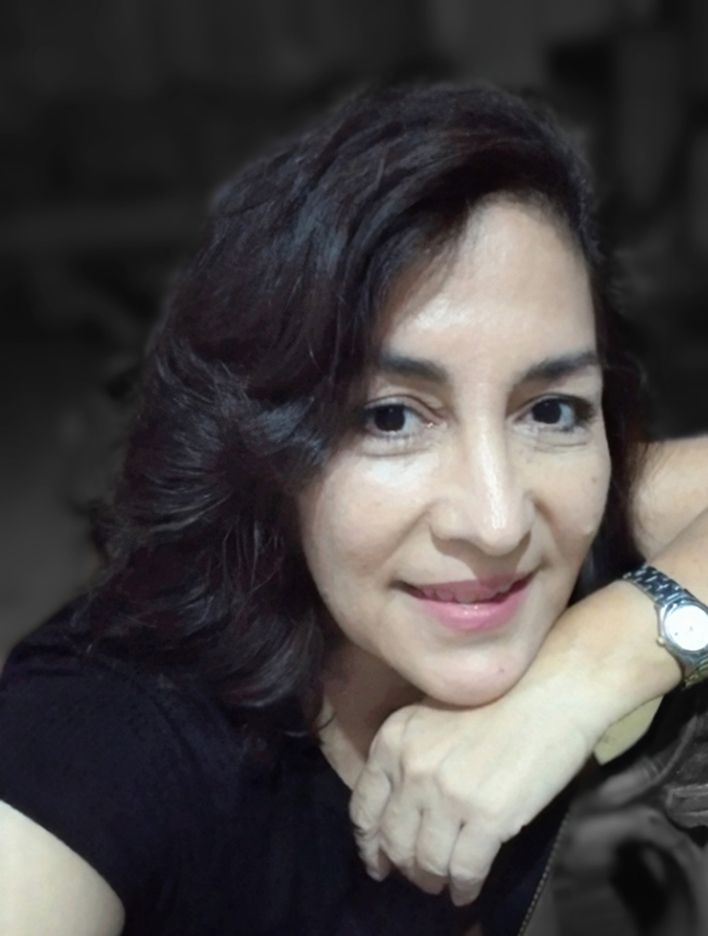 Woman with dark wavy hair smiles, resting arm on a dark surface. Wearing a watch and black shirt, neutral background.
