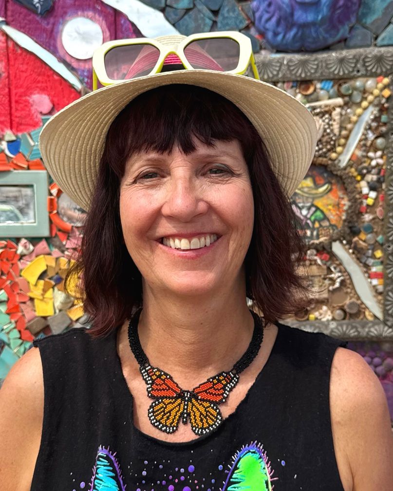 Woman in hat and sunglasses smiles, wearing a butterfly necklace, in front of mosaic art.