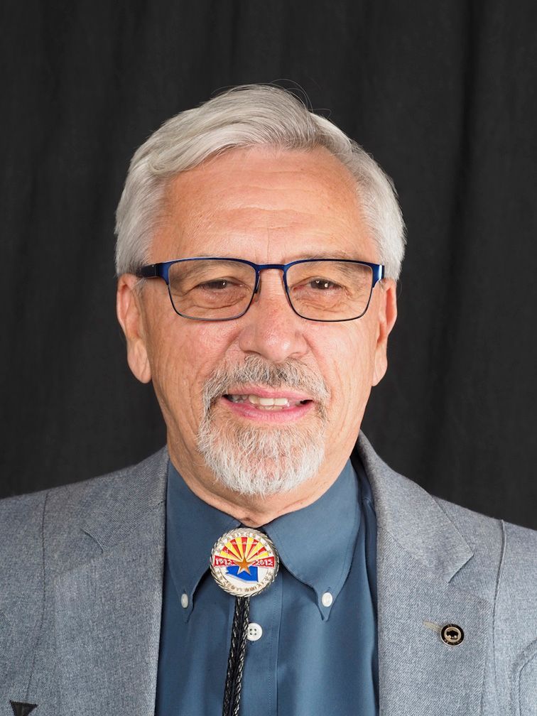 Man wearing glasses, gray blazer, and blue shirt with a bolo tie featuring the Arizona state seal.