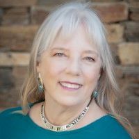 Woman with long, white hair smiles at the camera. She wears a turquoise top and silver necklace; brick background.